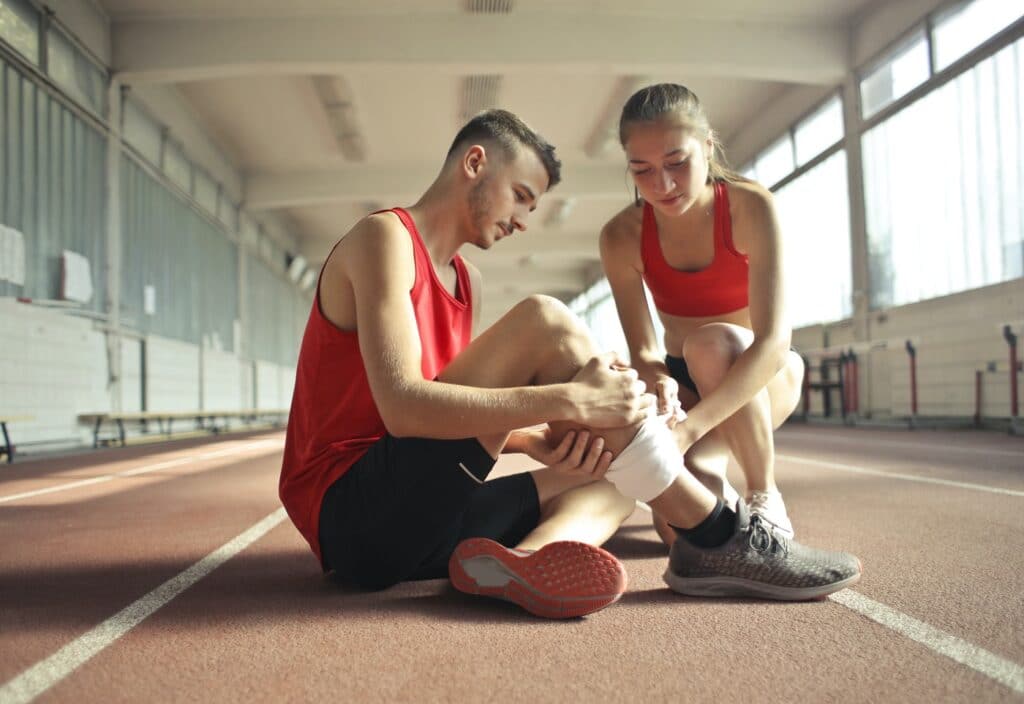 Two athletes on an indoor track with a female athlete wrapping the injured male athlete's leg to help treat his apparent sports injury
