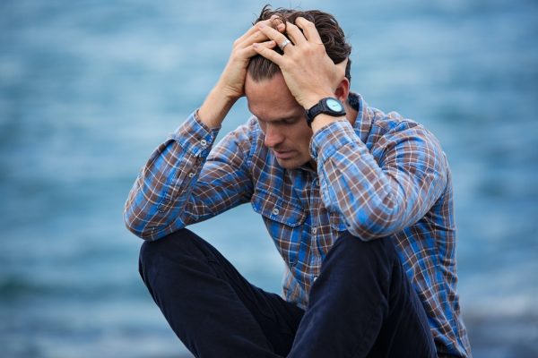 Man in a blue plaid shirt sitting with head in hands due to headaches or pain or frustration with water behind him